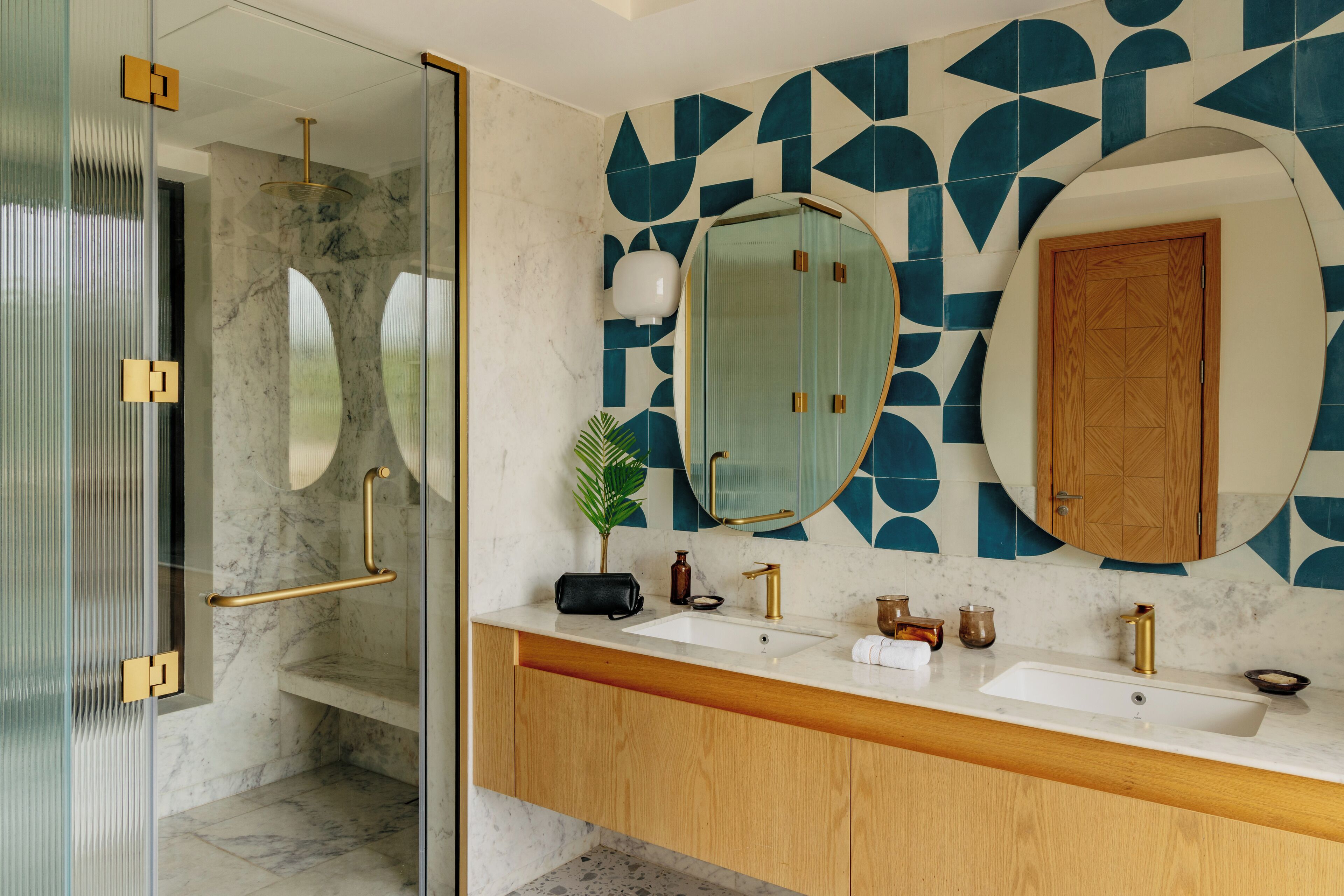 Un baño moderno con cabina de ducha de vidrio, pared con azulejos de mármol y tocador de madera con lavamanos blanco y detalles dorados.