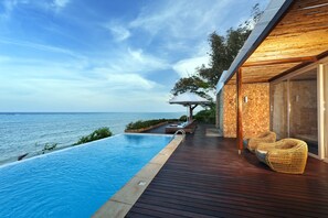 A wooden deck with a pool overlooking the sea, lounge chairs, and a stone wall.