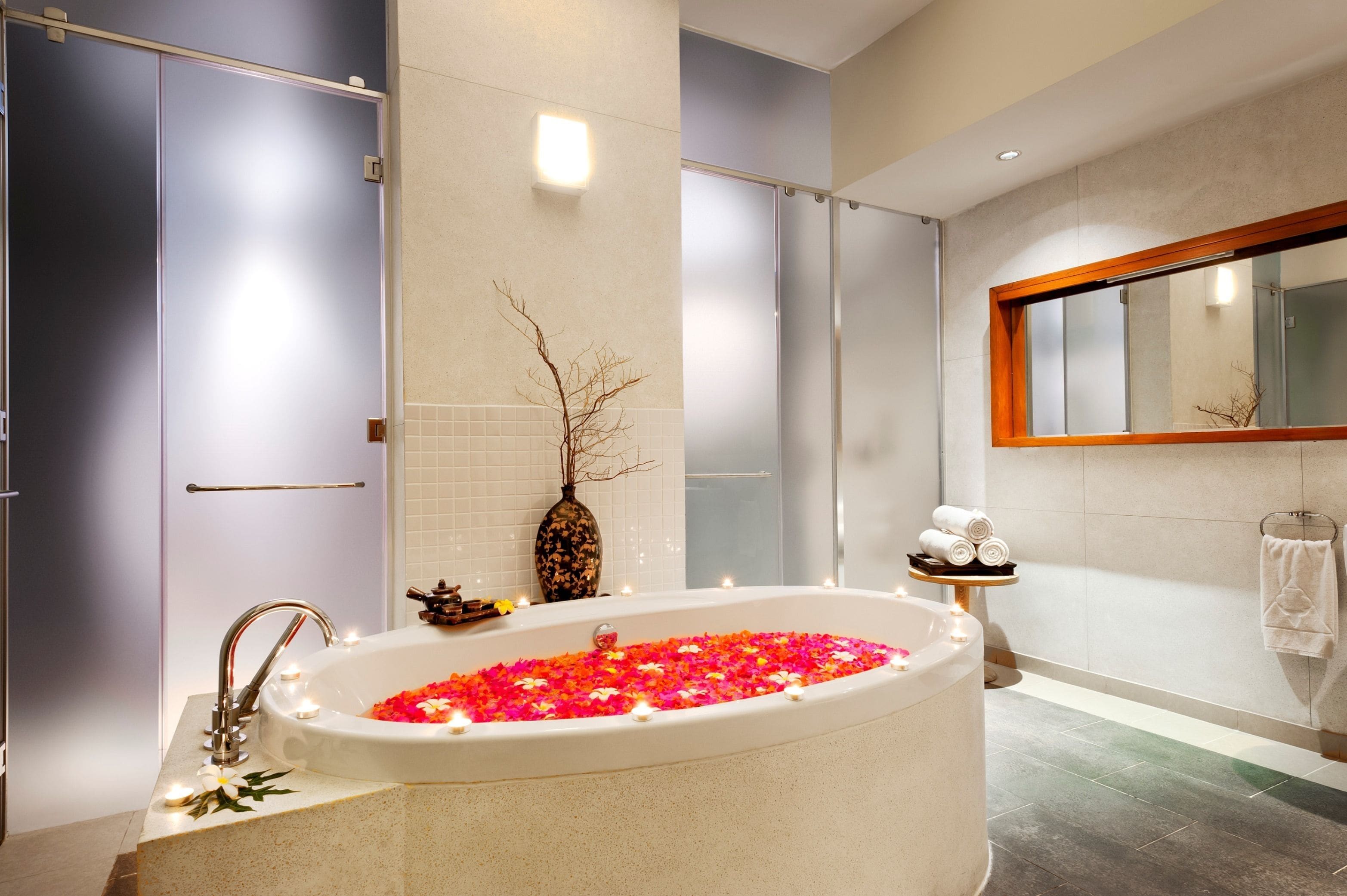 Un baño moderno con una bañera independiente llena de pétalos de rosa y velas, un espejo grande y un perchero.