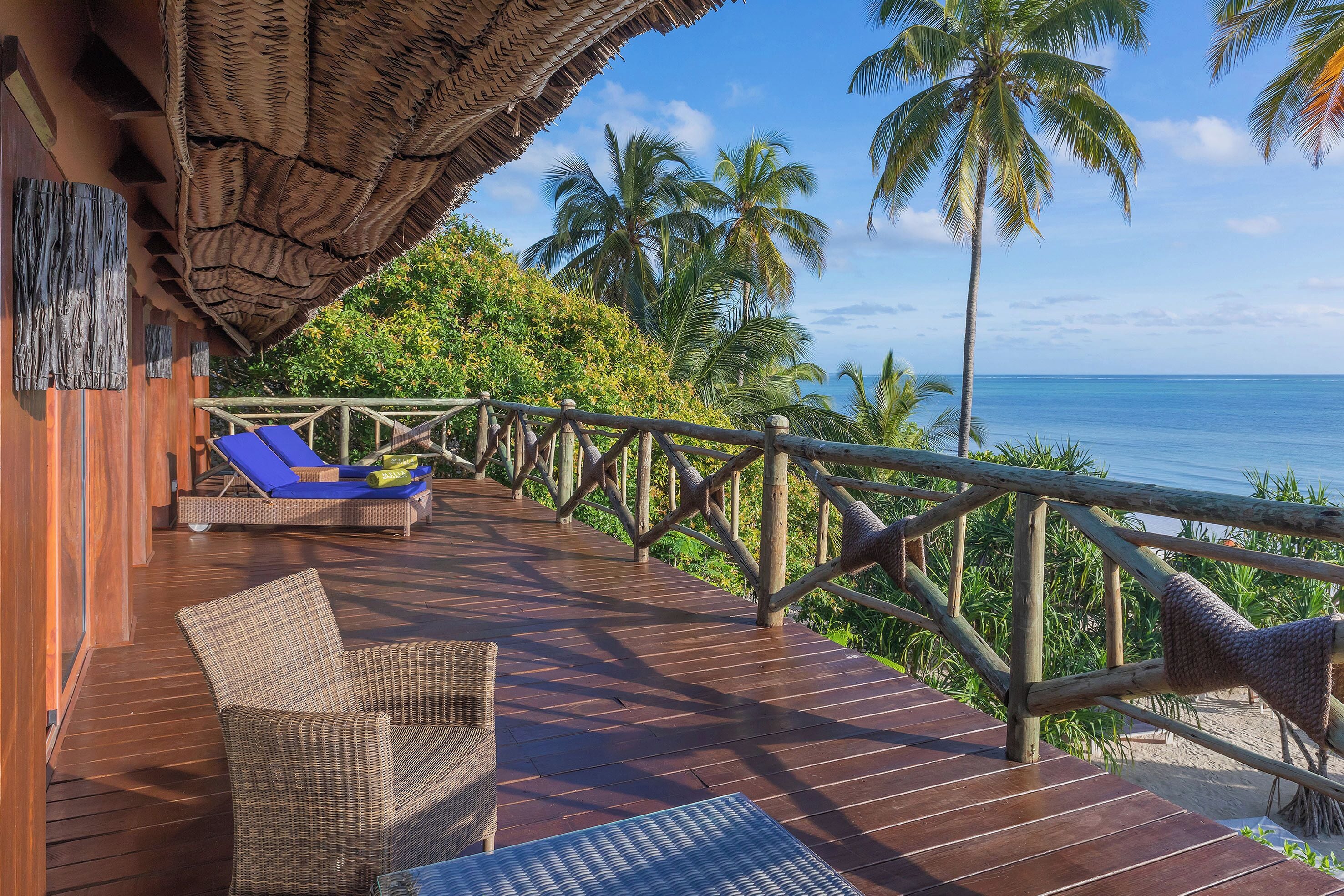 A wooden deck with a thatched roof, a wicker chair, a lounge chair, and a view of the ocean and palm trees.