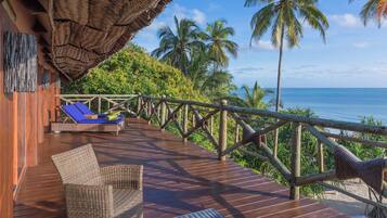 A wooden deck with a thatched roof, a wicker chair, a lounge chair, and a view of the ocean and palm trees.