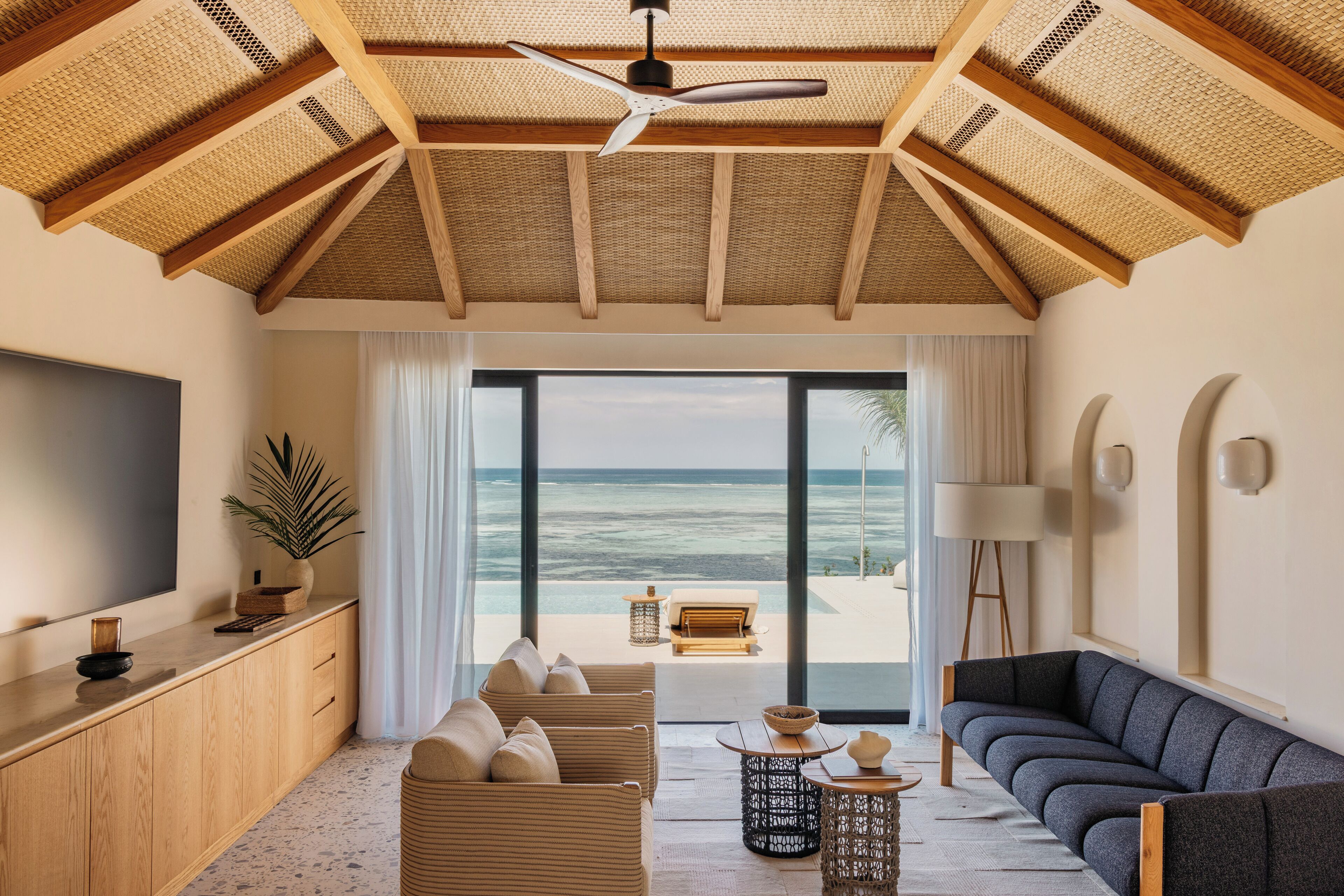 Una sala de estar moderna con un gran ventanal con vistas al océano, un ventilador de techo y un televisor a la izquierda.