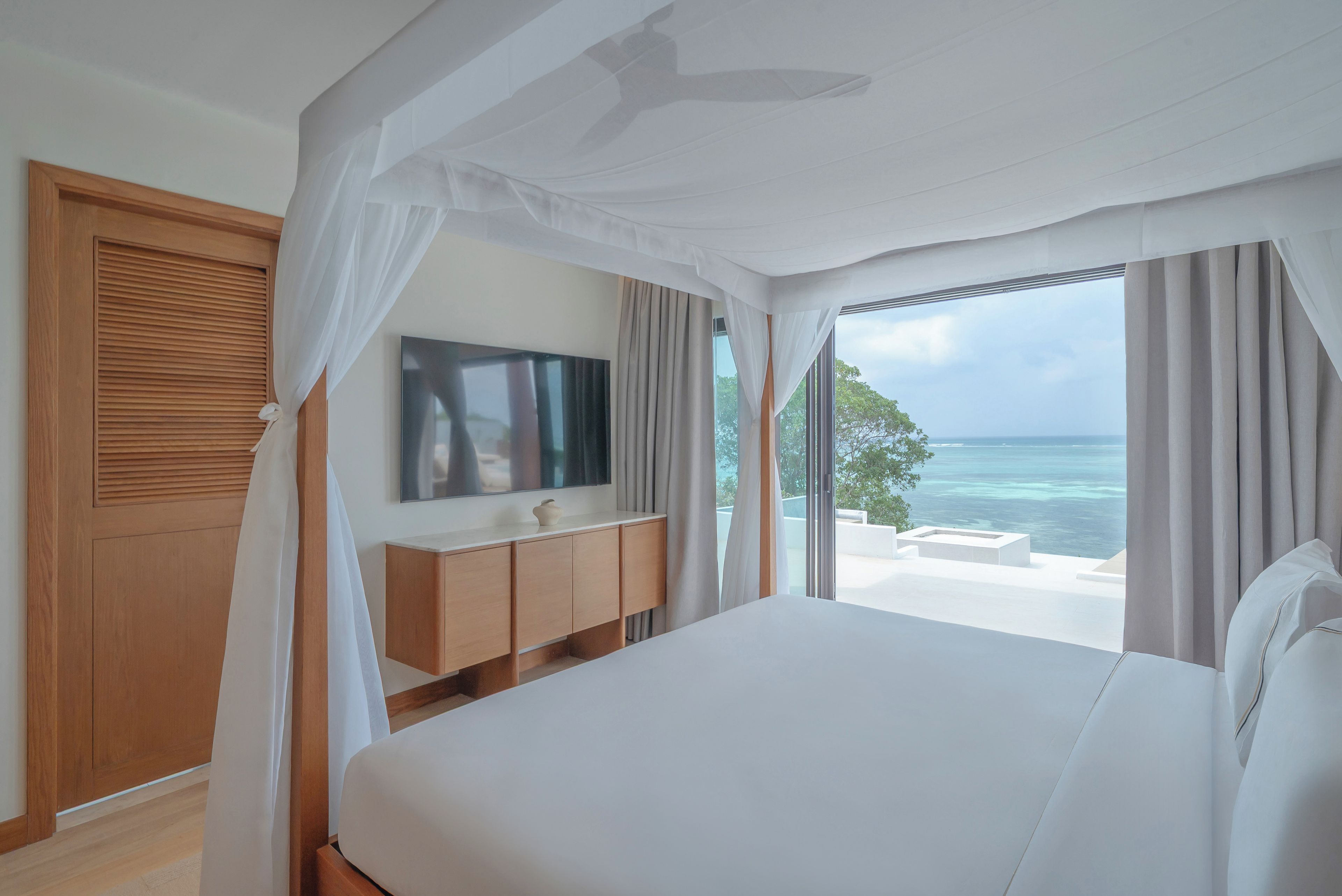 A bedroom with a large bed, a TV on a wooden cabinet, and a view of the ocean.