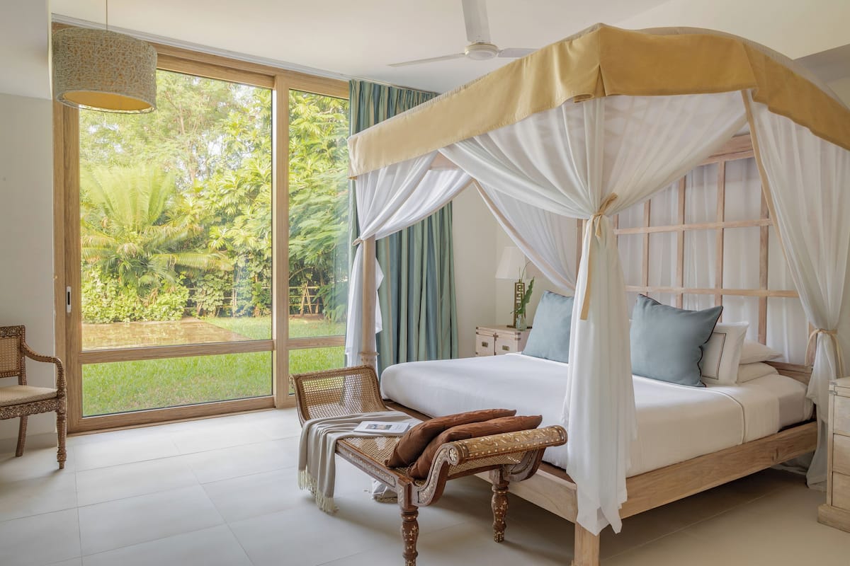 Ein geräumiges Schlafzimmer mit einem großen Bett, einer Sitzbank und Blick auf einen üppigen Garten durch eine Schiebetür.
