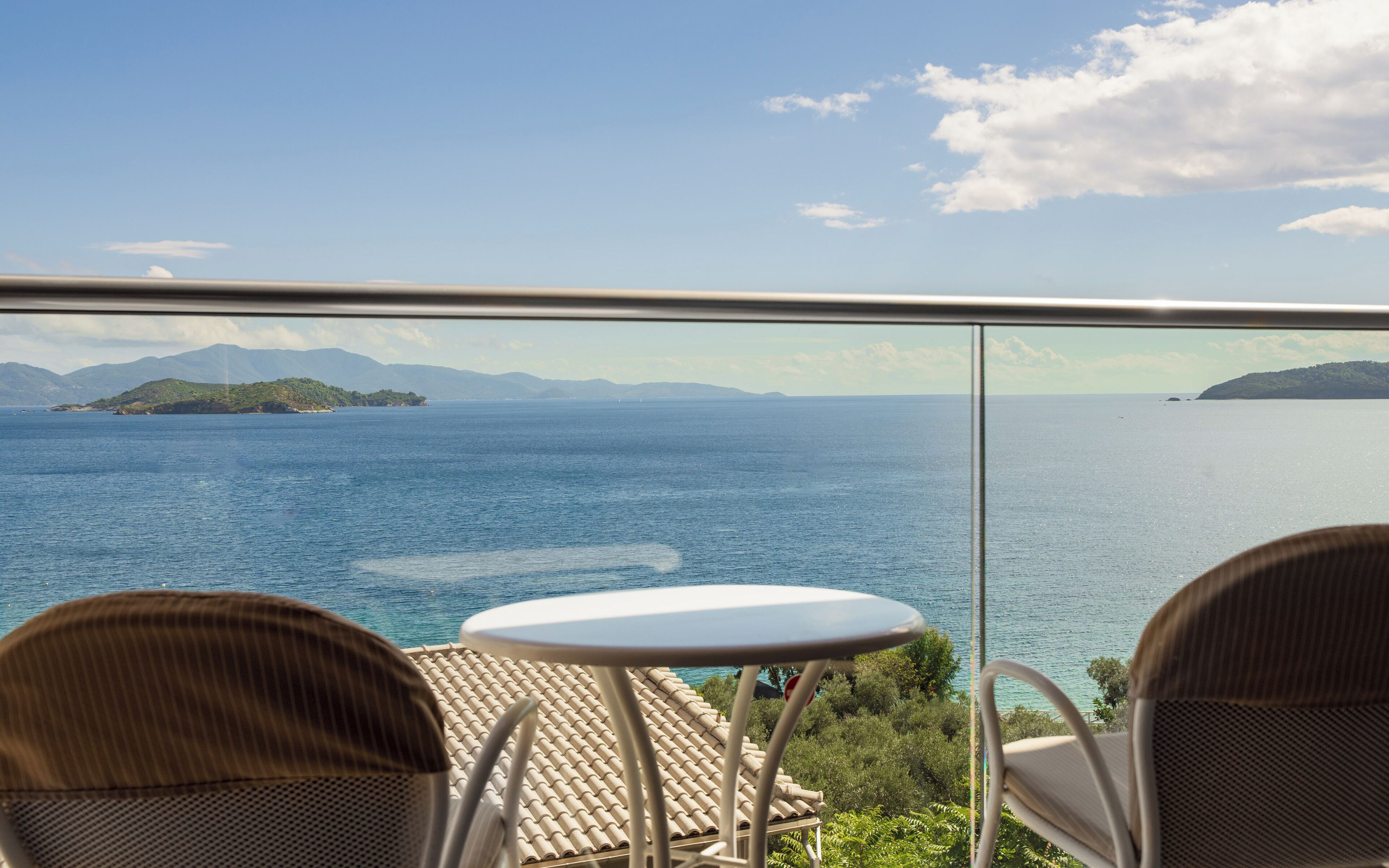 A balcony with two wicker chairs and a small table, overlooking a calm sea and distant islands.