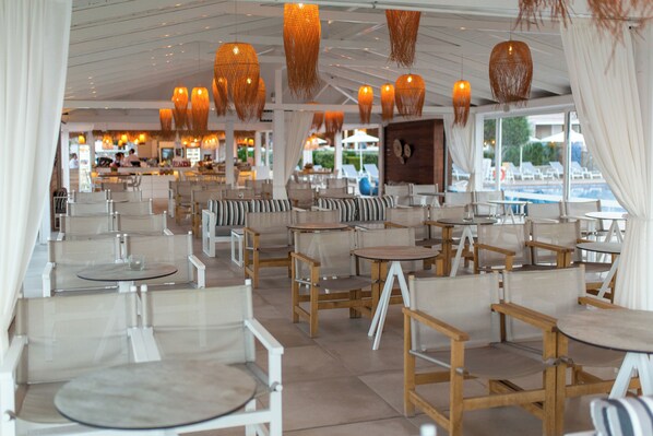 Un restaurante con una decoración moderna, que cuenta con mesas y sillas de madera, lámparas de mimbre colgantes y grandes ventanales con vistas a una zona de piscina.