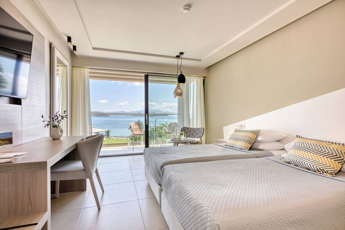 A modern hotel room with a large bed, a desk, and a balcony with a view of the sea.
