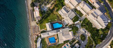 Vista aérea de un complejo turístico costero con varias edificaciones, una playa y un estacionamiento.