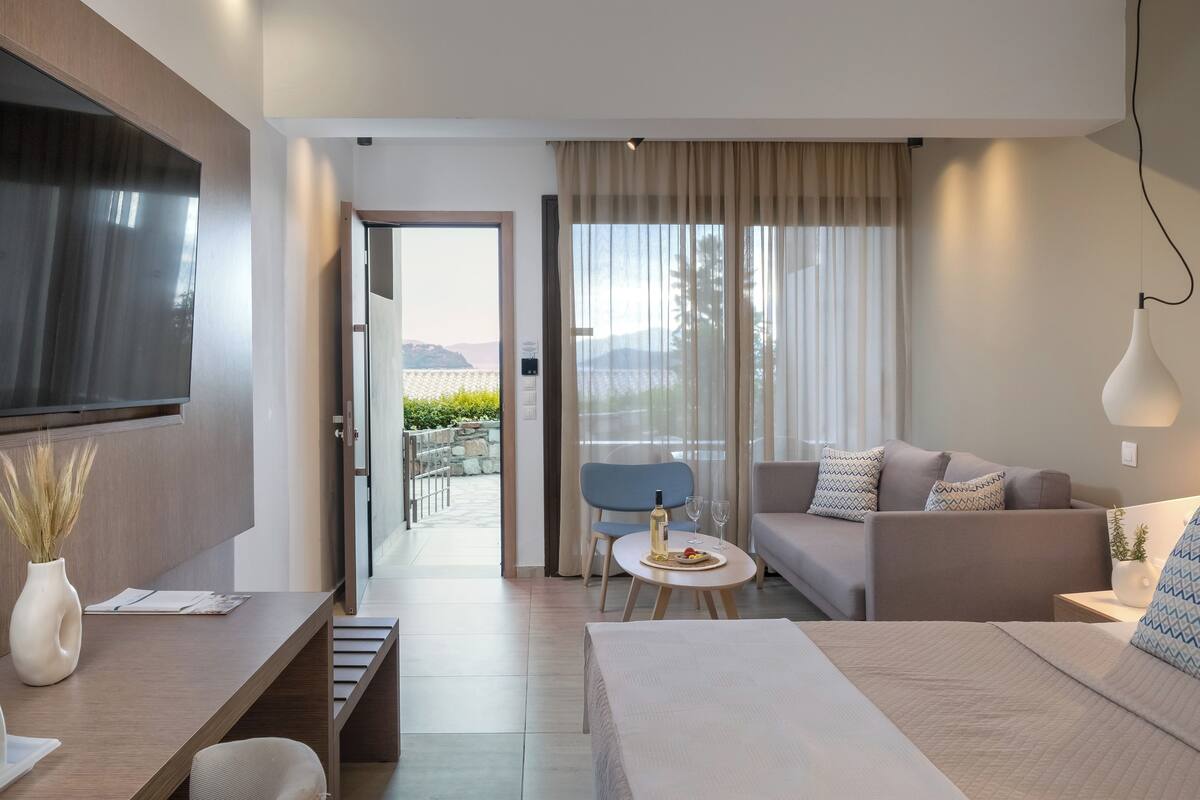 A modern hotel room with a bed, a sofa, a TV, a dining table with a bottle and glasses, and a view of the outdoors.