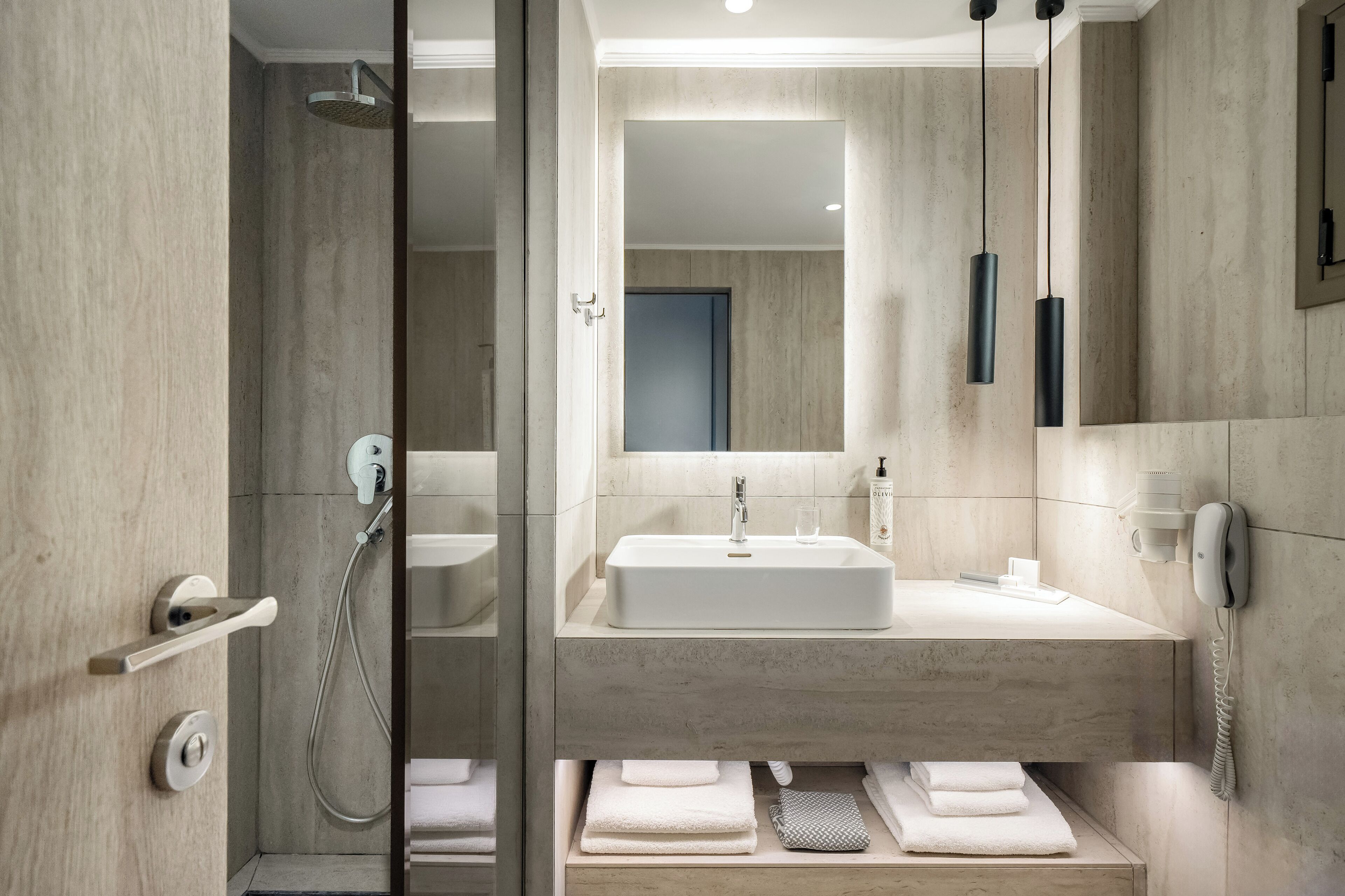 Un baño moderno con ducha envuelta en vidrio, un espejo grande, un lavamanos blanco y un perchero con toallas dobladas con cuidado.