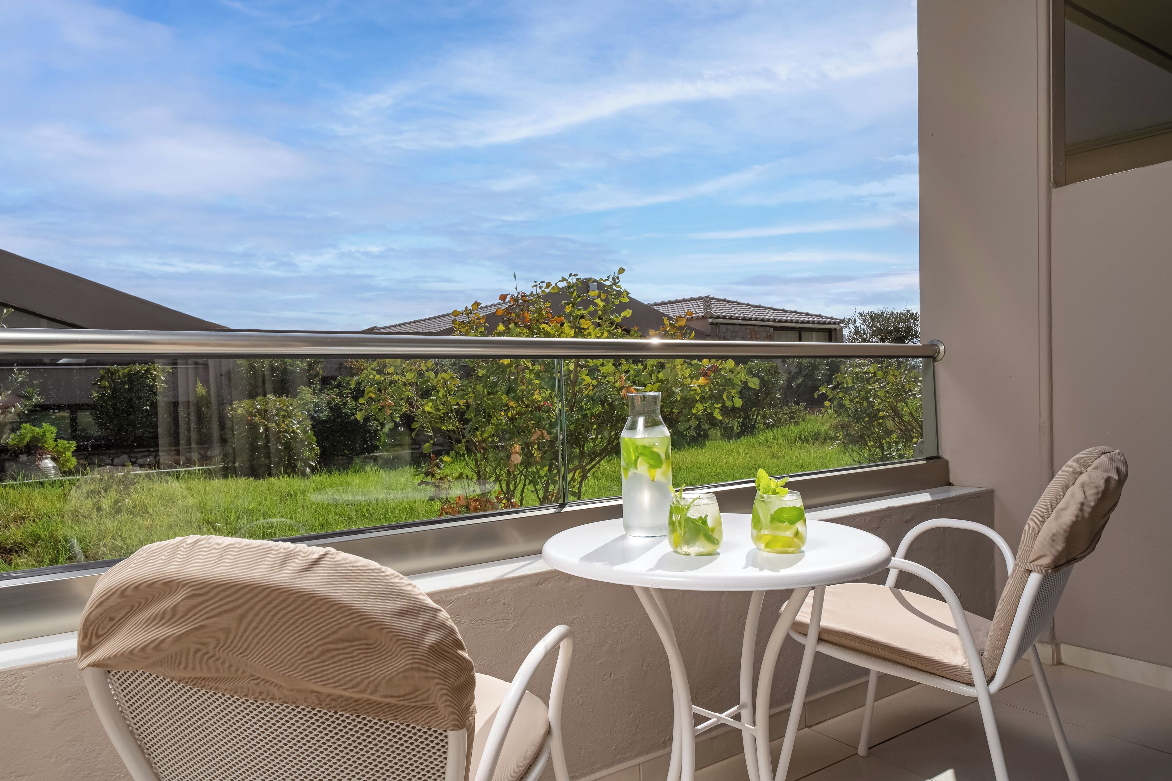A balcony with a white table and chairs, a clear view of a garden, and a bottle of water.