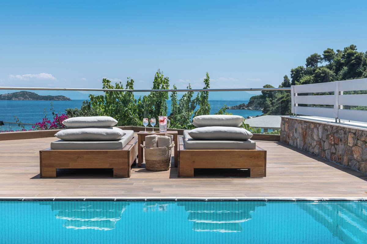 A poolside area with wooden loungers, a stone wall, and a view of the sea.