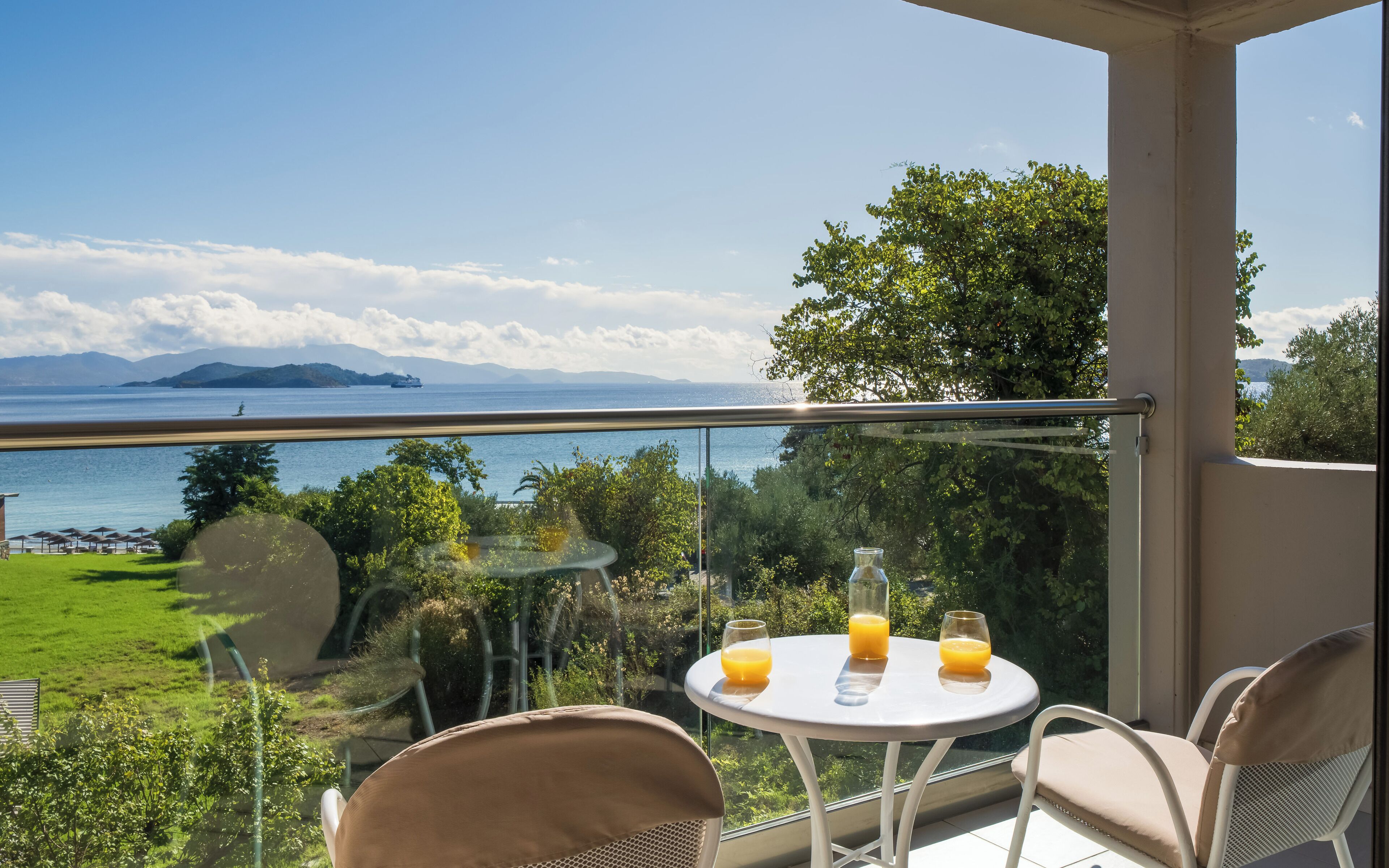 A balcony with a glass railing overlooking a sea view, two chairs, a small table, and two glasses of orange juice.