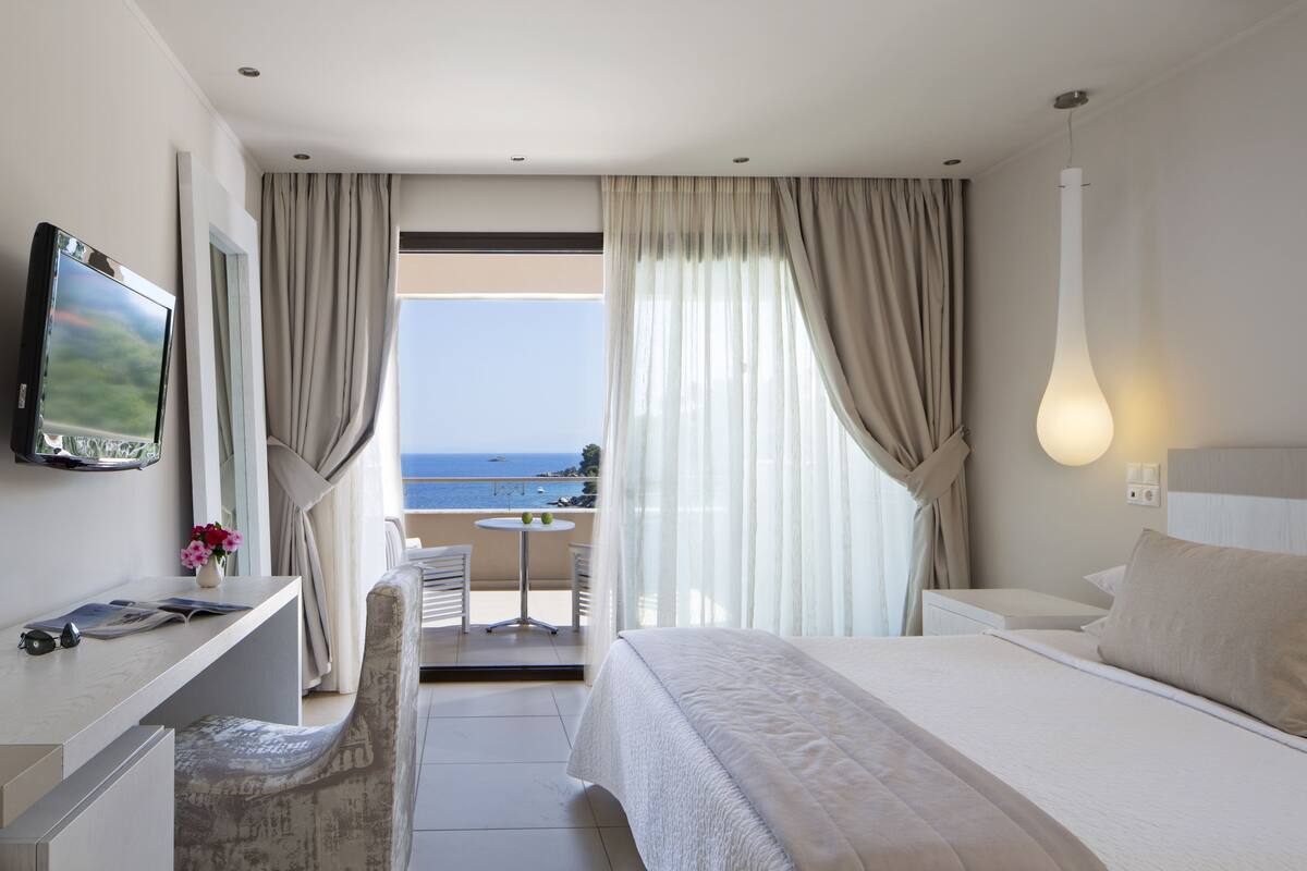 A modern hotel room with a bed, a desk with a TV, a small table with chairs, and a balcony with a view of the sea.