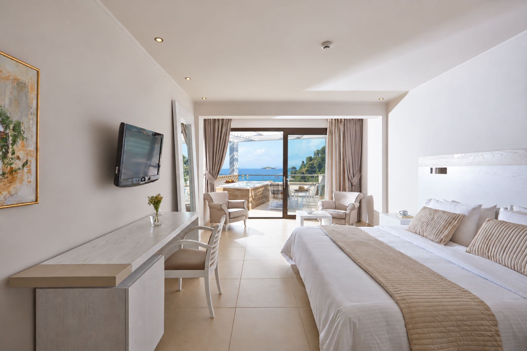 A hotel room with a bed, a desk, a chair, a TV, and a balcony with a view of the sea.