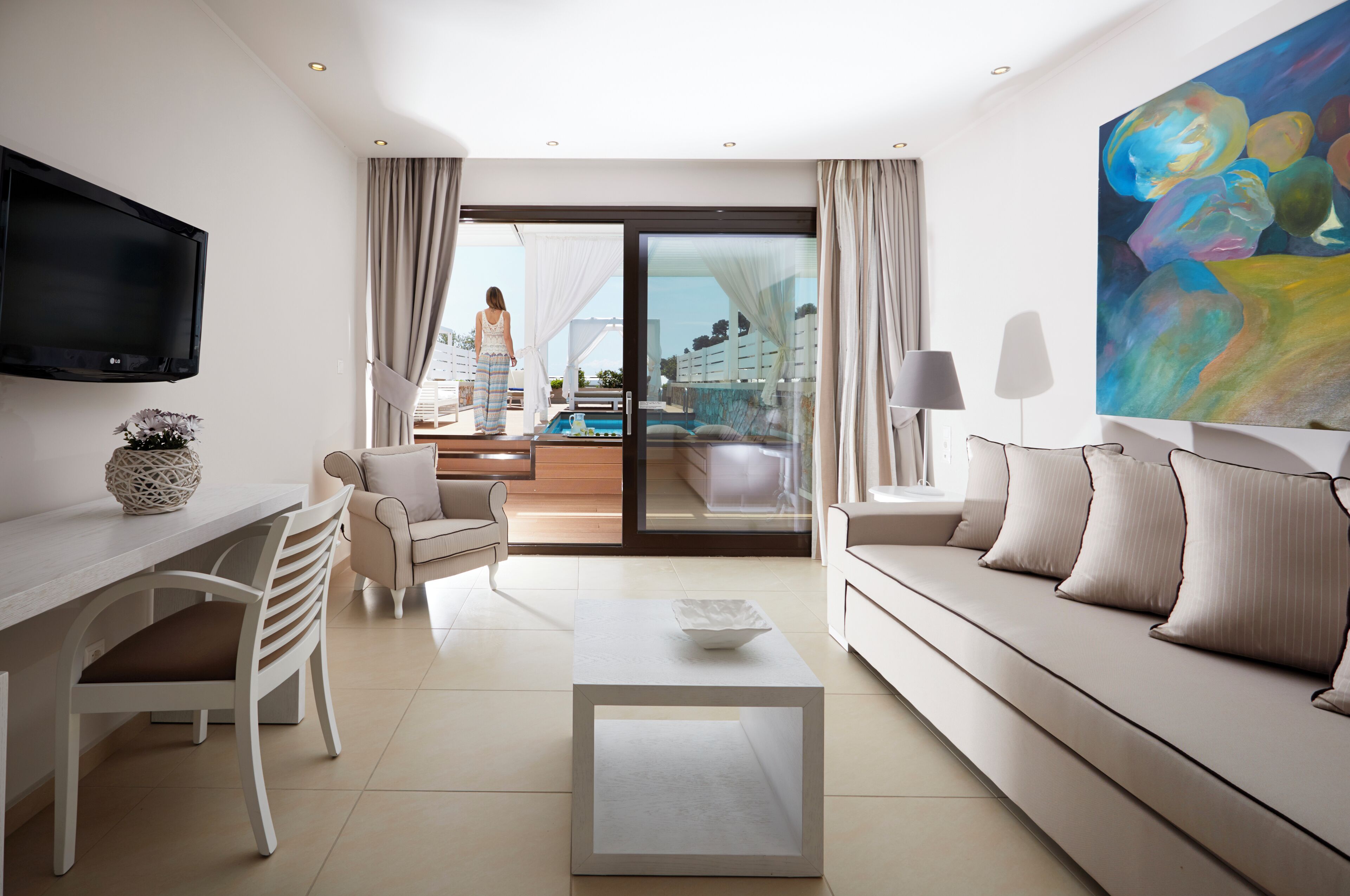 A modern hotel room with a sofa, a coffee table, a TV, and a view of a pool and balcony.