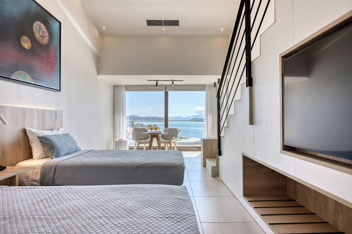A modern hotel room with two beds, a large TV, a balcony with a view of the sea, and a staircase.