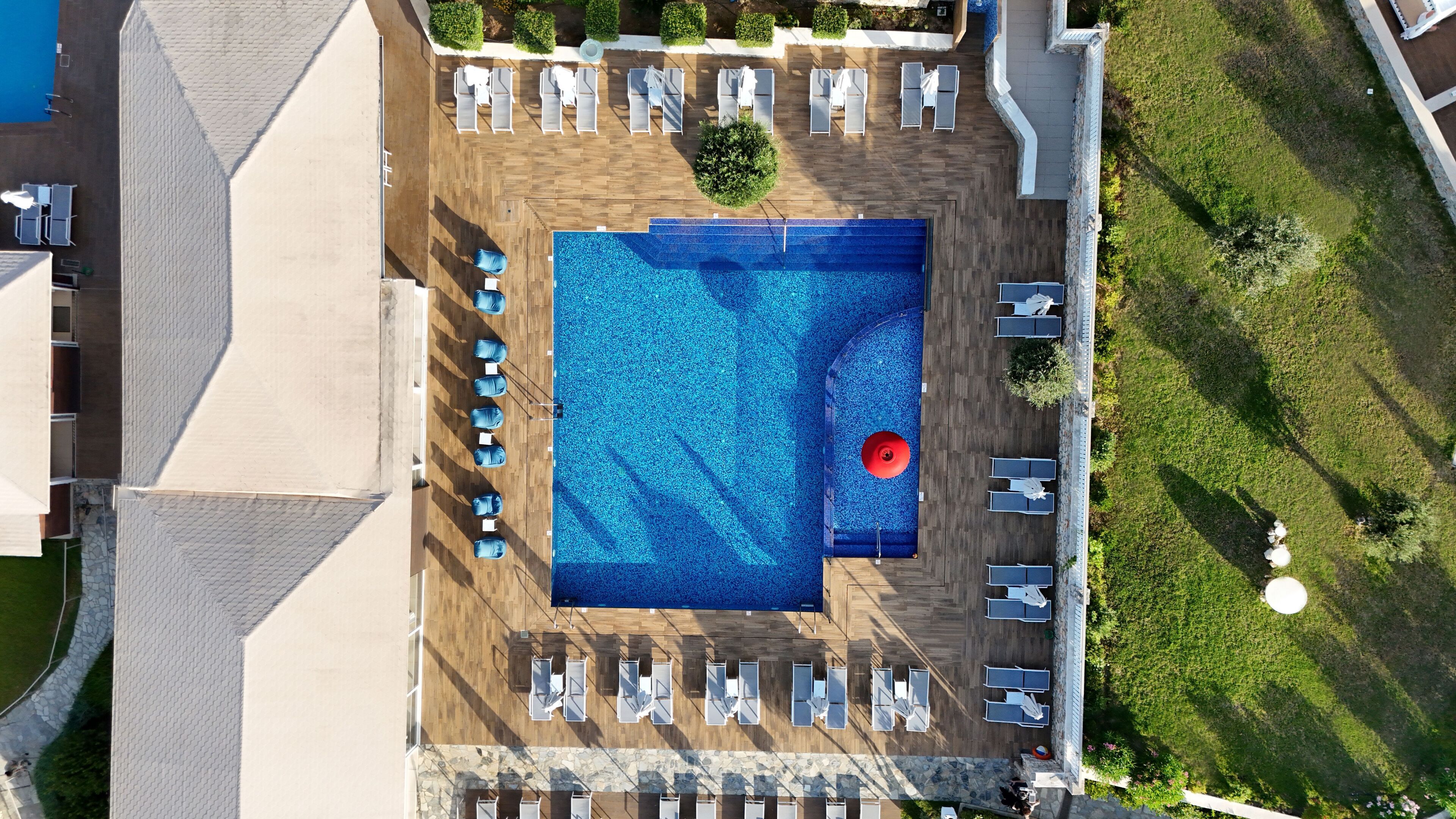 An aerial view of a hotel with a large swimming pool surrounded by sun loungers and a small red circular feature, possibly a hot tub or fountain.