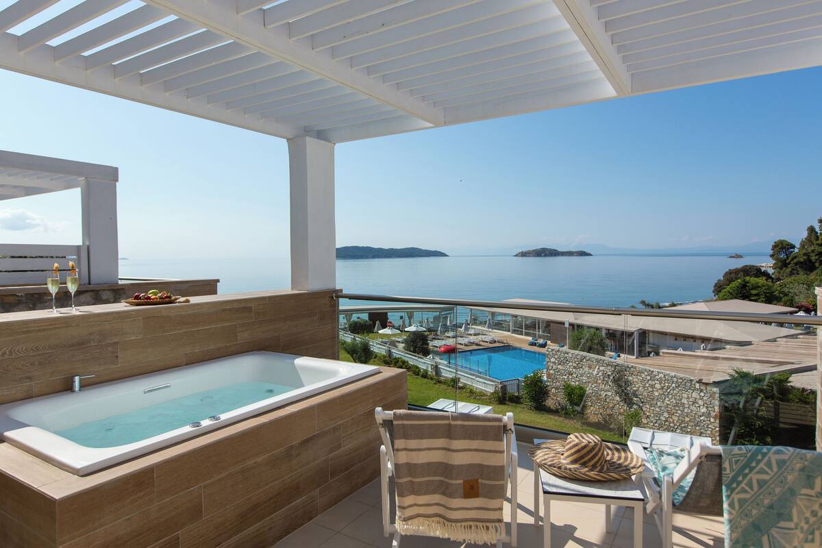 A balcony with a hot tub, outdoor seating, and a view of a swimming pool and the sea.