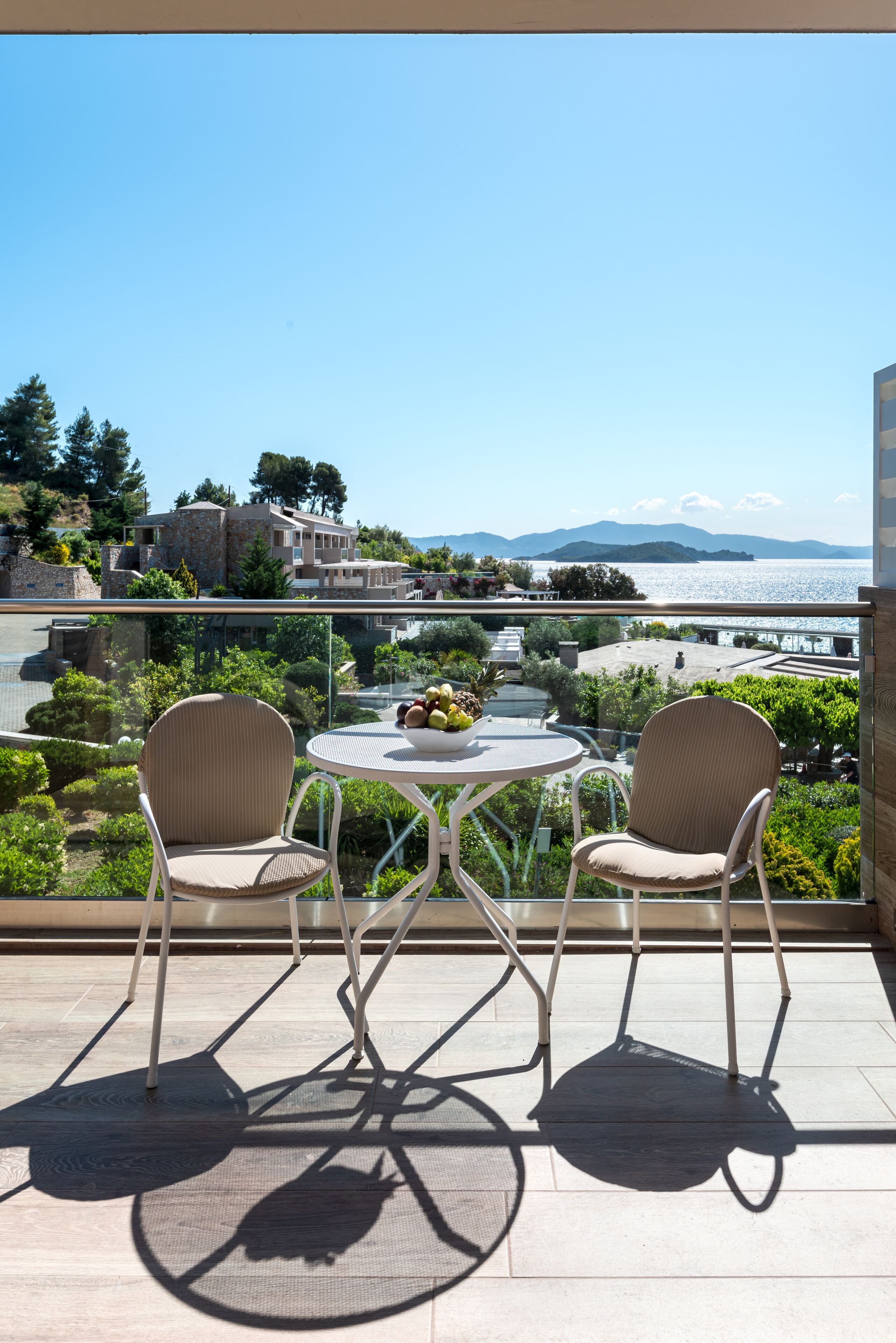 A balcony with two wicker chairs and a small round table, overlooking a scenic view of a coastal landscape with buildings and mountains in the distance.