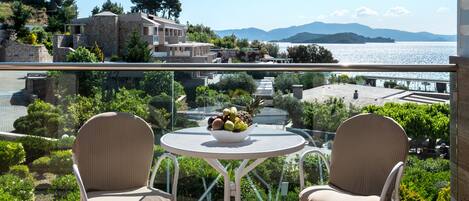 A balcony with two wicker chairs and a small round table, overlooking a scenic view of a coastal landscape with buildings and mountains in the distance.