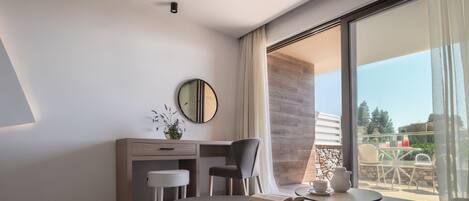 A modern hotel room with a large window, a desk, a chair, a small table, and a coffee table.