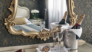 A luxurious room with a large ornate mirror, a bed with a blue and white bedspread, a bedside table with a vase of white flowers, and a tray with champagne bottles and glasses. - Hotel Antica Locanda Al Gambero (Venice)