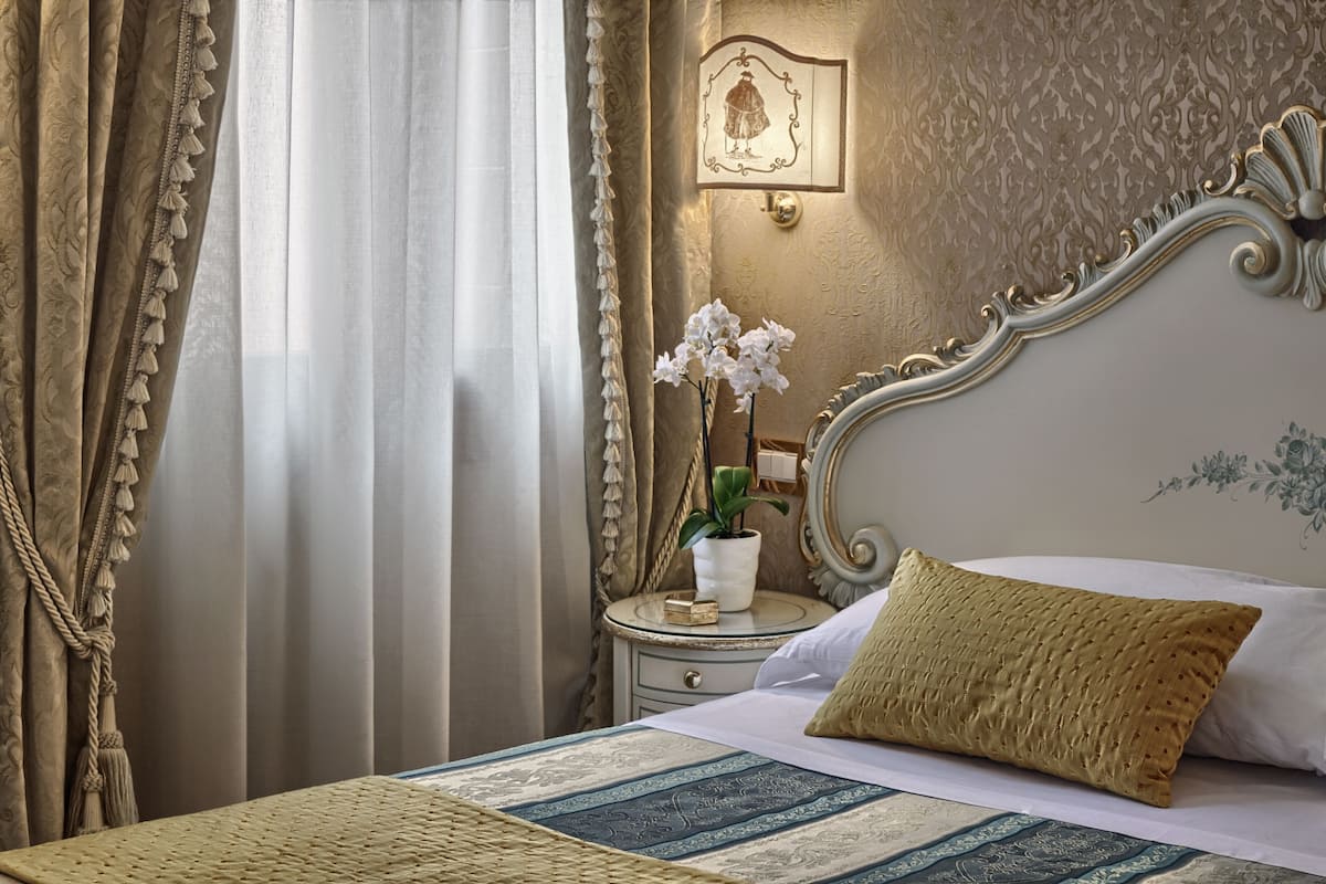 Een net opgemaakt bed met een bedsprei met patroon, een nachtkastje met een potplant, en een aan de muur bevestigde fotolijst.