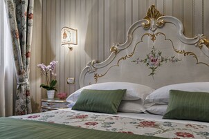 A hotel room with a bed, floral bedspread, green pillows, a nightstand with a lamp, and a wall-mounted light fixture.