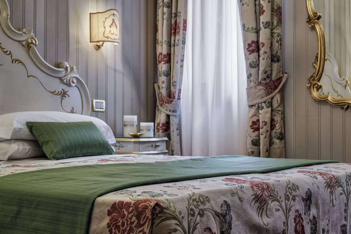 Een net opgemaakt bed met een bloemenbedekking en een groen kussen, een nachtkastje met een lamp, en gordijnen met een bloemenprint.