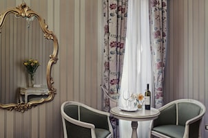Una stanza con un ampio specchio decorato, due poltrone imbottite, un tavolino con una bottiglia di vino, bicchieri e fiori.