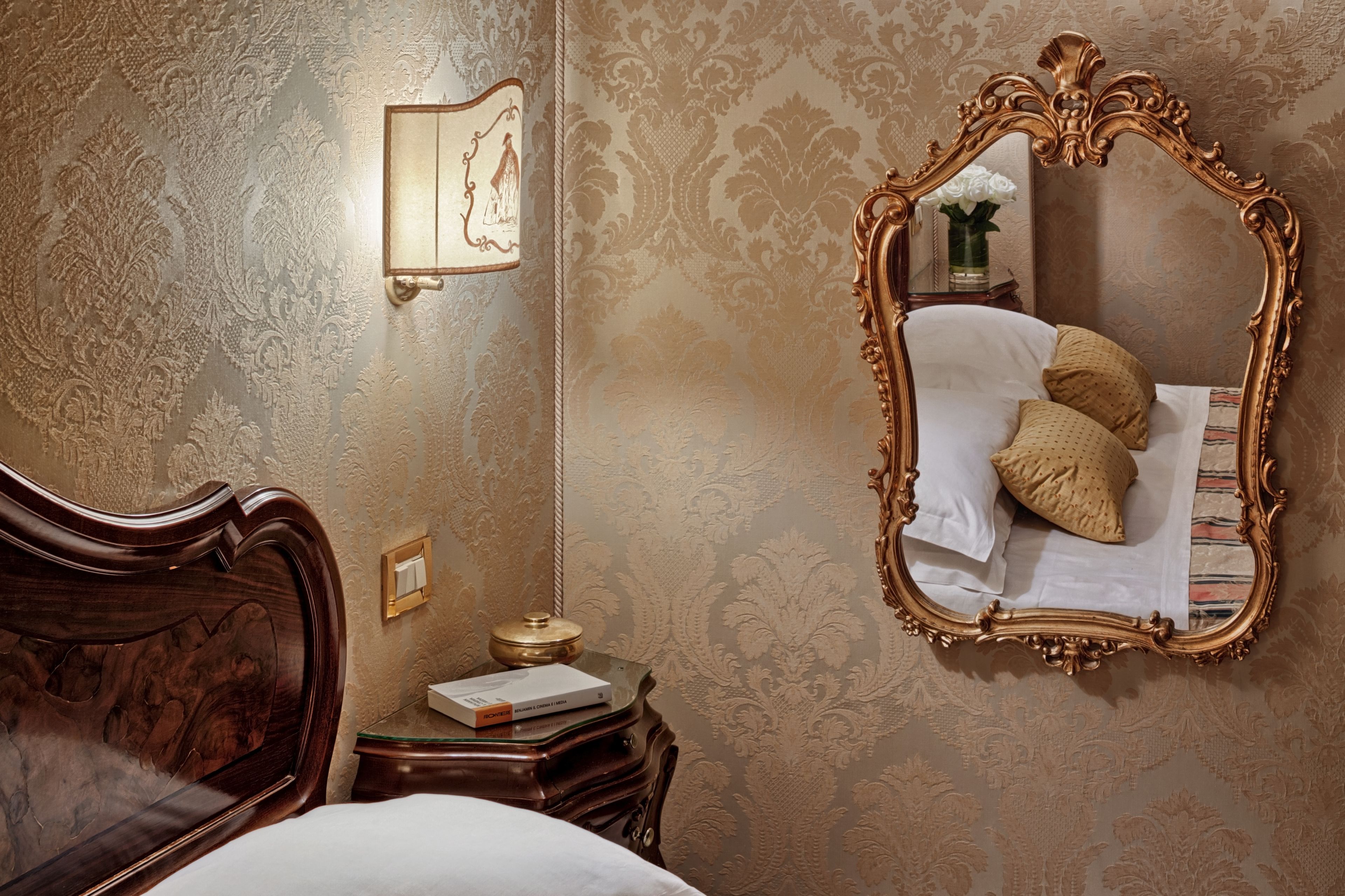 A bedroom with a large ornate mirror, a wooden headboard, a bedside table with a lamp, and a wall-mounted light fixture.