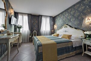 Ein Hotelzimmer mit einem großen Bett, einem Schreibtisch, einem Sessel, einem Spiegel und einem an der Wand befestigten Fernseher.