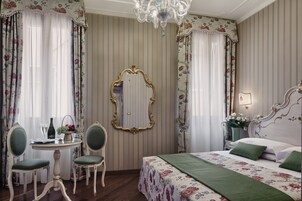 A bedroom with a bed, two chairs, a small table, a mirror, and a chandelier.