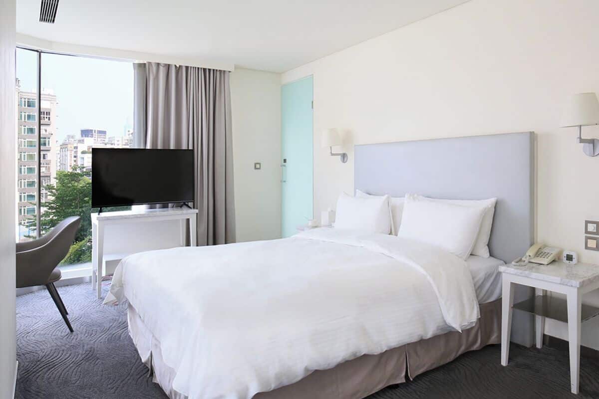 Een hotelkamer met een groot bed, een tv op een standaard, een nachtkastje met telefoon, en een stoel bij het raam.
