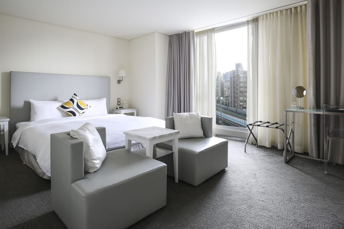 Een moderne hotelkamer met een groot bed, twee fauteuils, een bijzettafeltje en uitzicht op een stadsgezicht door grote ramen.