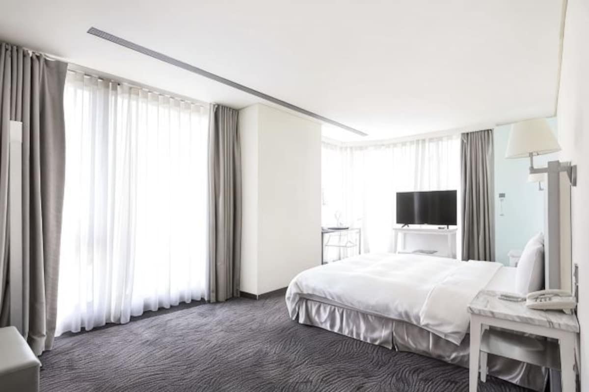 A hotel room with a large bed, a desk with a lamp, a TV, and a window with curtains.