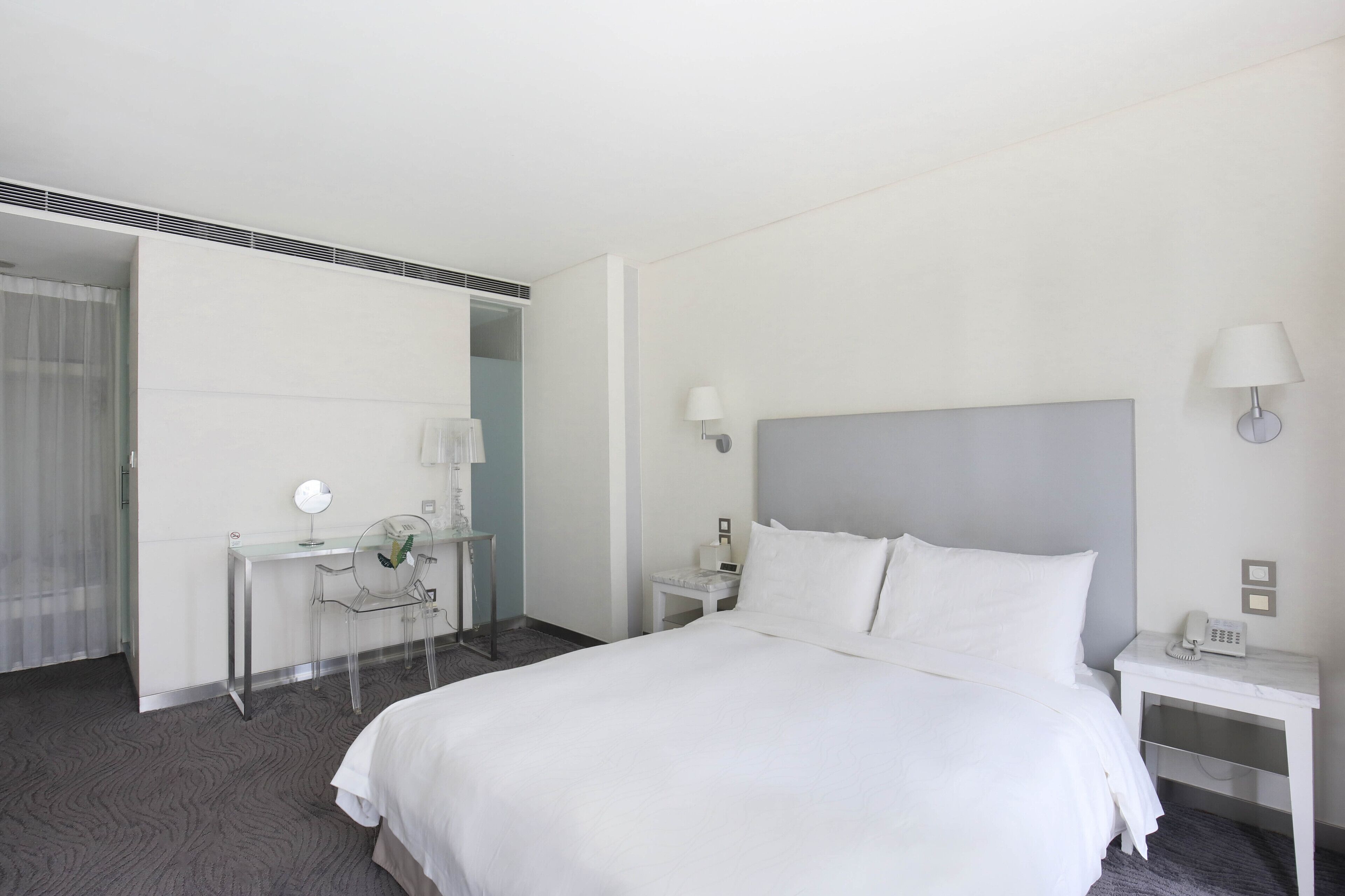 A hotel room with a large bed, a desk with a chair, a mirror, a lamp, and a phone on the nightstand.