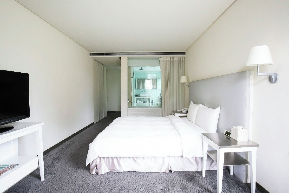 A hotel room with a bed, a TV on a stand, a bedside table with a lamp, and a view of a bathroom through a glass door.