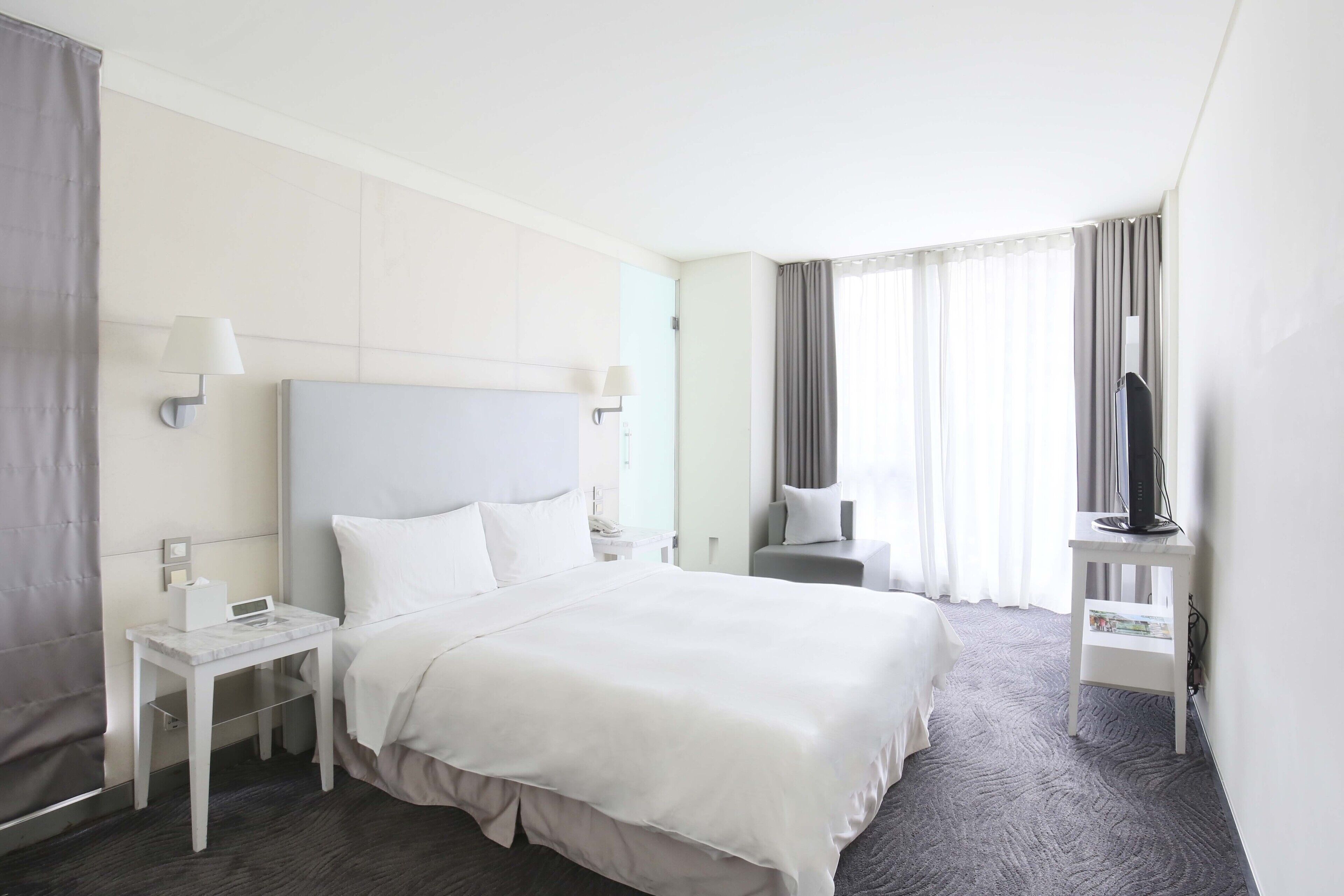 Quarto de hotel com uma cama grande, mesa de cabeceira, televisão sobre um suporte e janela com cortinas.