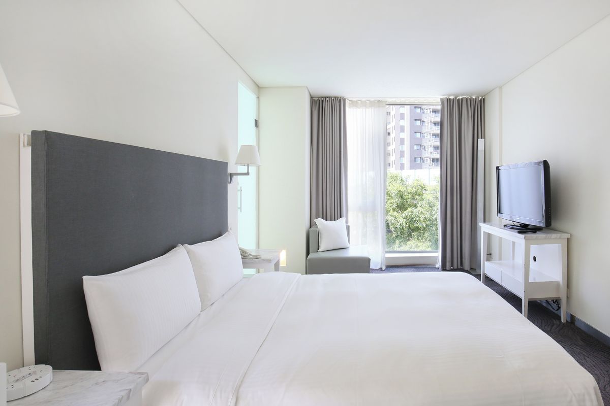 A hotel room with a large bed, a television on a stand, a window with curtains, and a view of buildings outside.