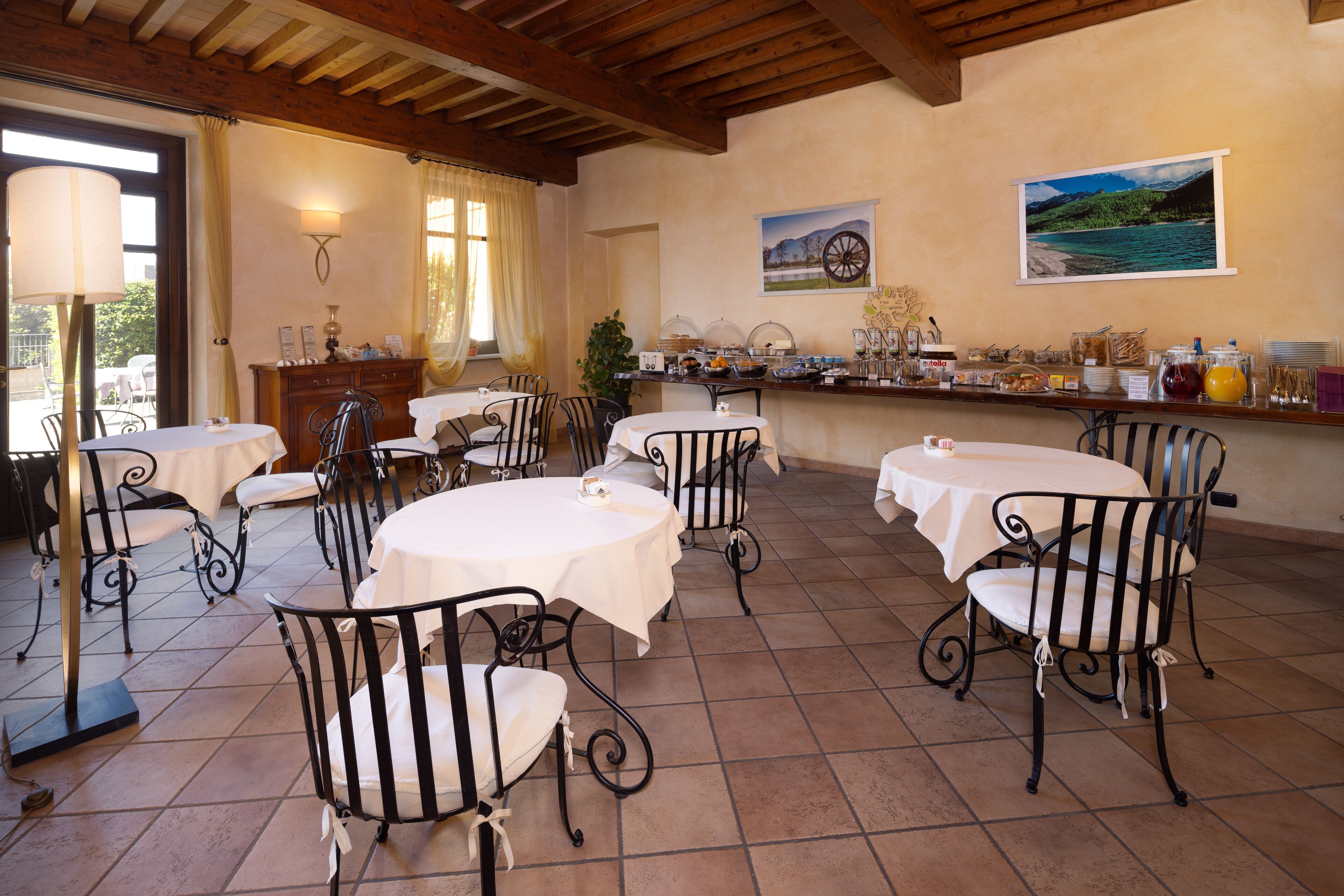 A dining area with round tables and metal chairs, a buffet with various food items, and a scenic painting on the wall.