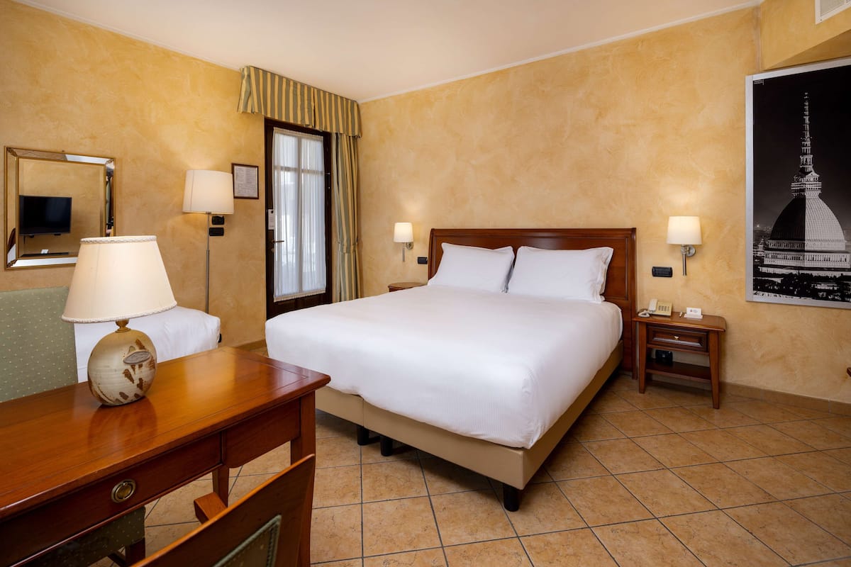 Une chambre d’hôtel avec un grand lit, deux tables de chevet avec des lampes, un bureau avec une lampe, une chaise, un miroir et un tableau encadré au mur.