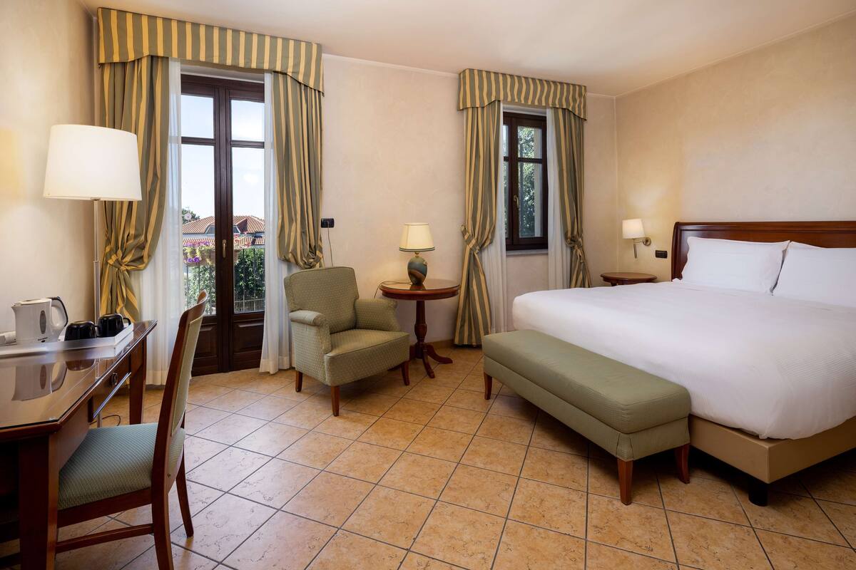 Une chambre d’hôtel avec un lit, un bureau avec une lampe, une chaise, une petite table avec une lampe et des rideaux.