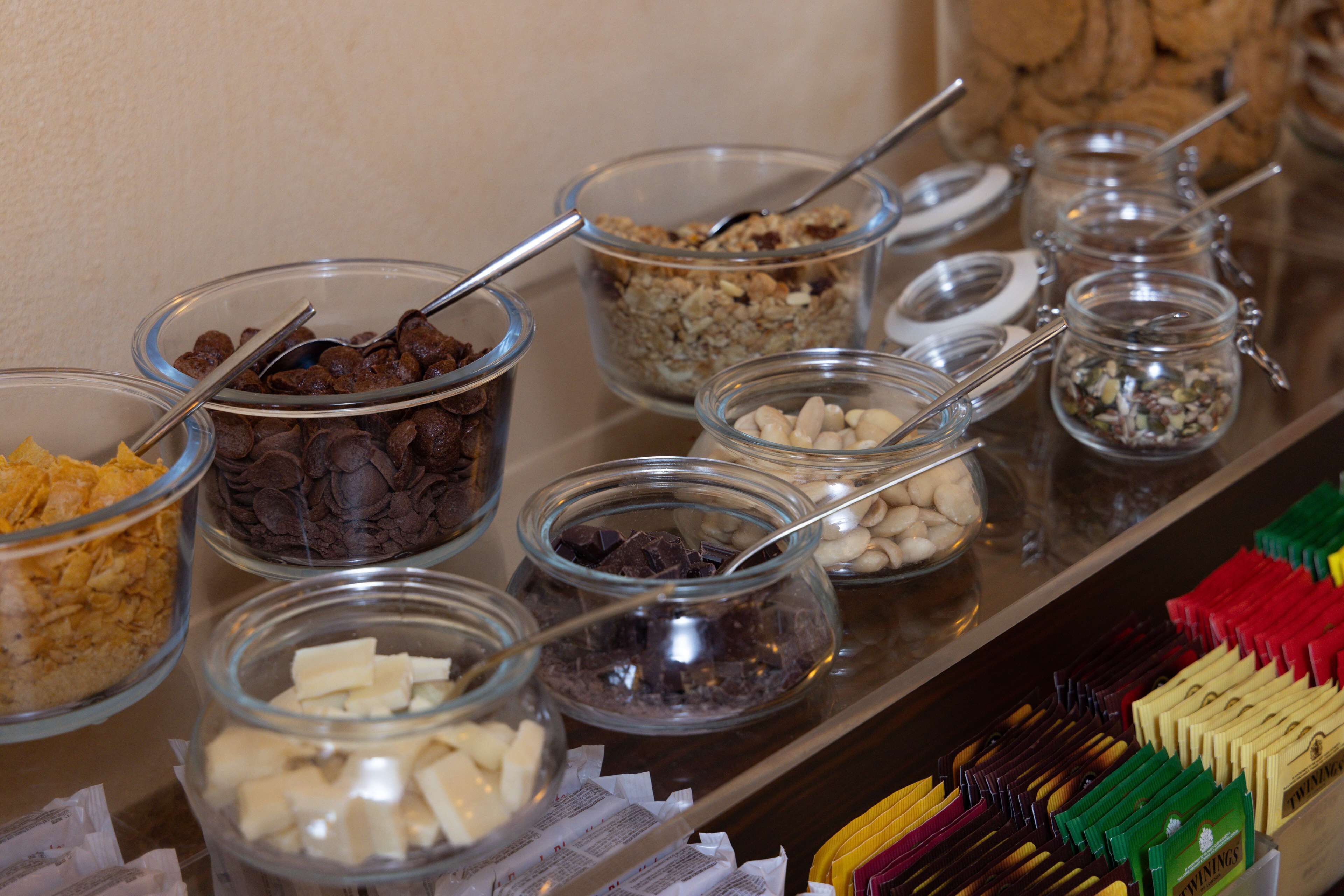 Un bufé con diversos alimentos en recipientes de vidrio, como cereales, frutos secos y trozos de chocolate.