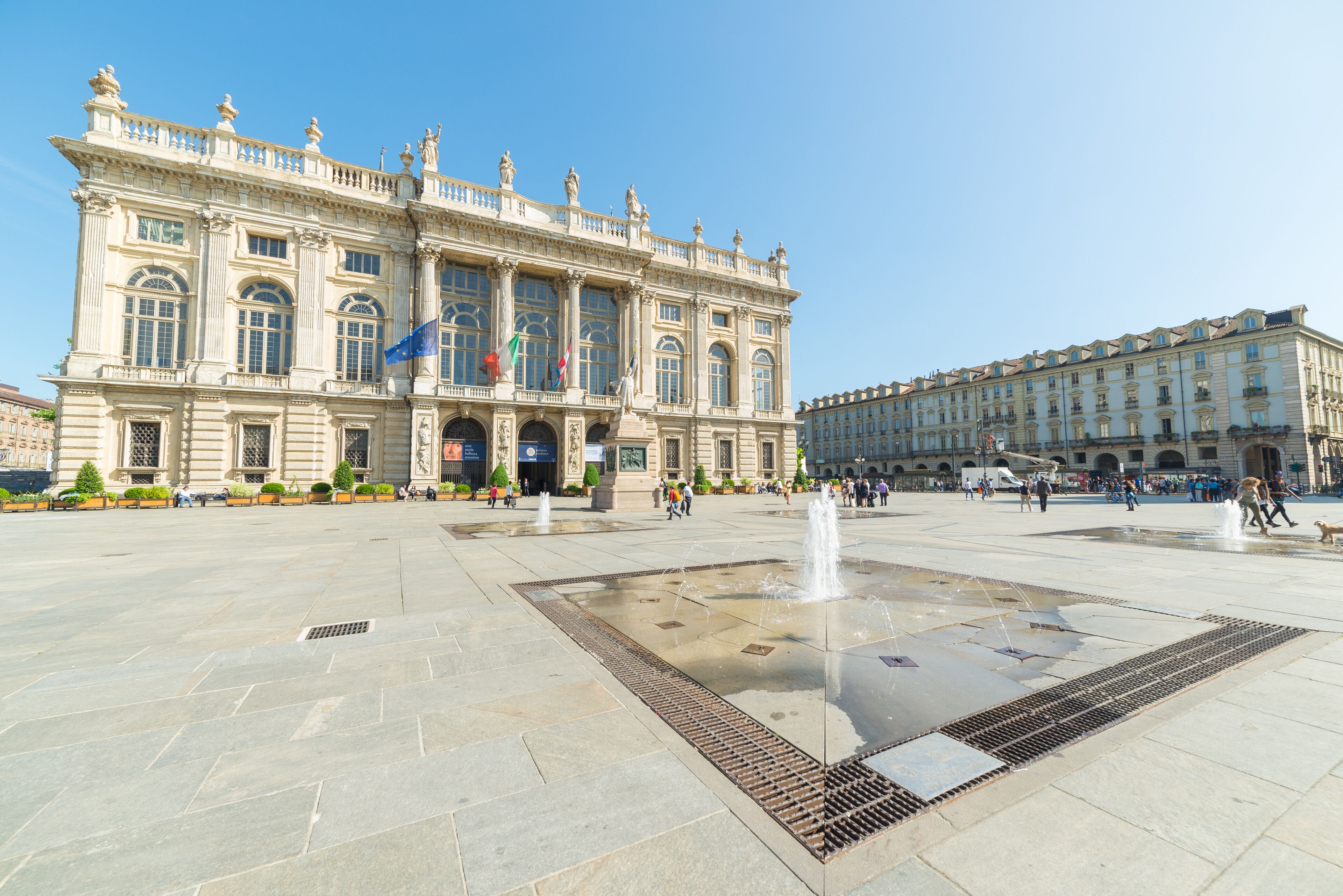 A large, historic building with classical architecture, a central fountain, and a spacious plaza.