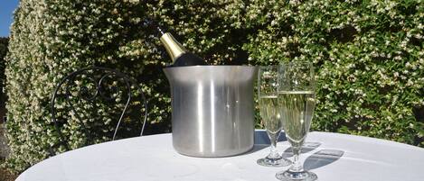 A table with a champagne bottle and two glasses, set outdoors with a hedge and a building in the background.