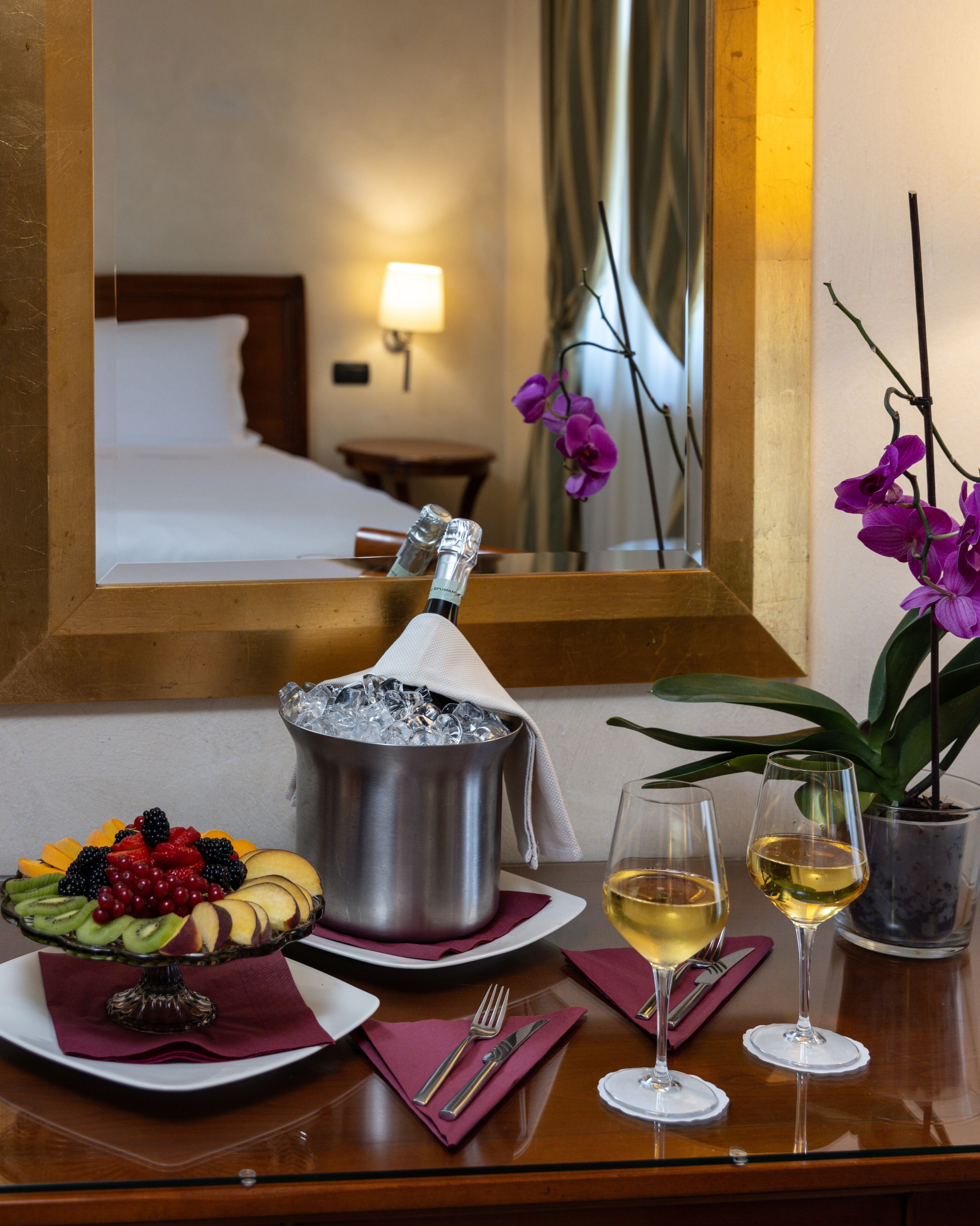 Une chambre d’hôtel avec un lit, une table de chevet sur laquelle se trouve un panier de fruits, une bouteille de champagne, deux verres à vin et une orchidée en pot.