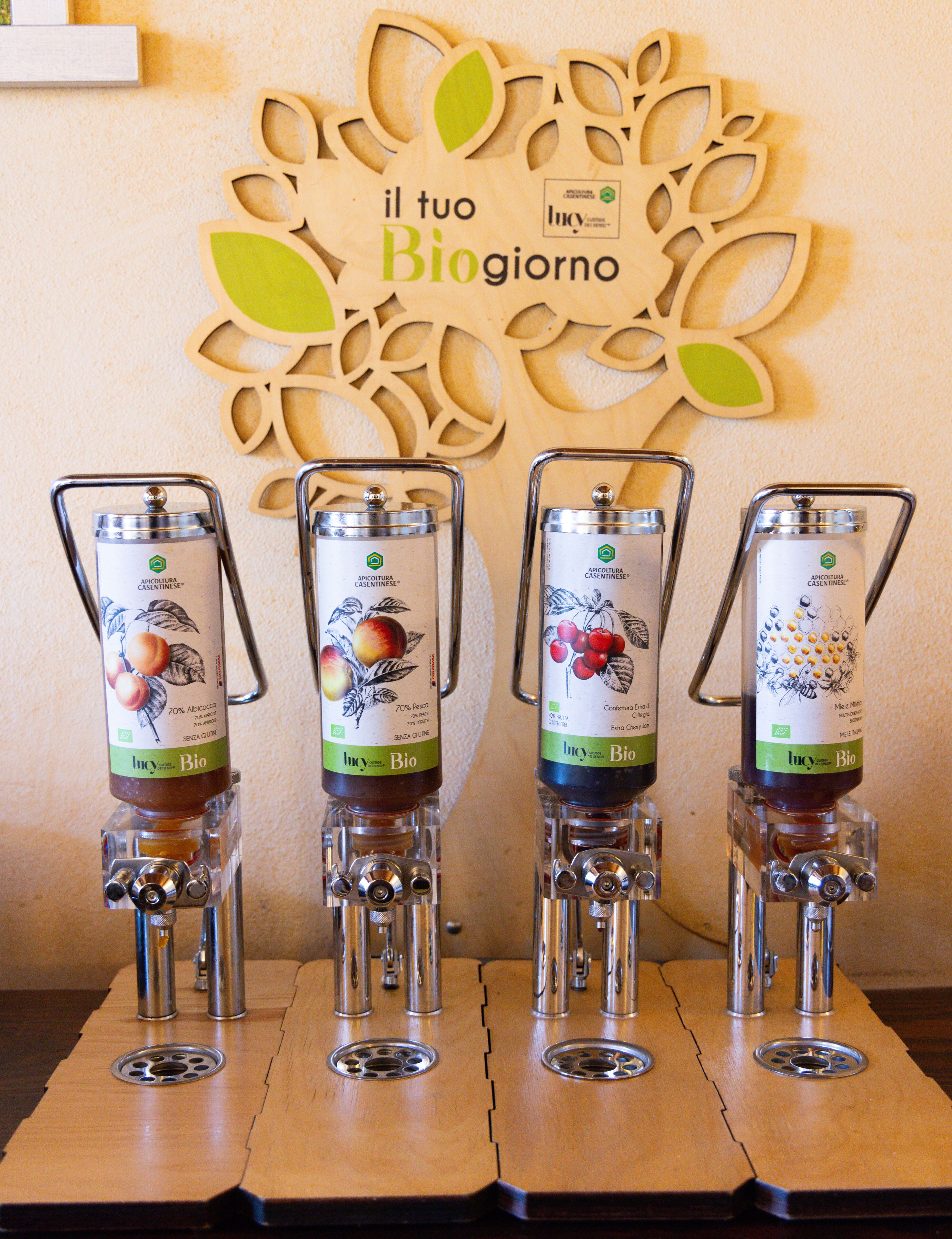 Four bottles of 'Lucy Bio' brand apple cider vinegar on a wooden counter with a decorative wall piece behind them.