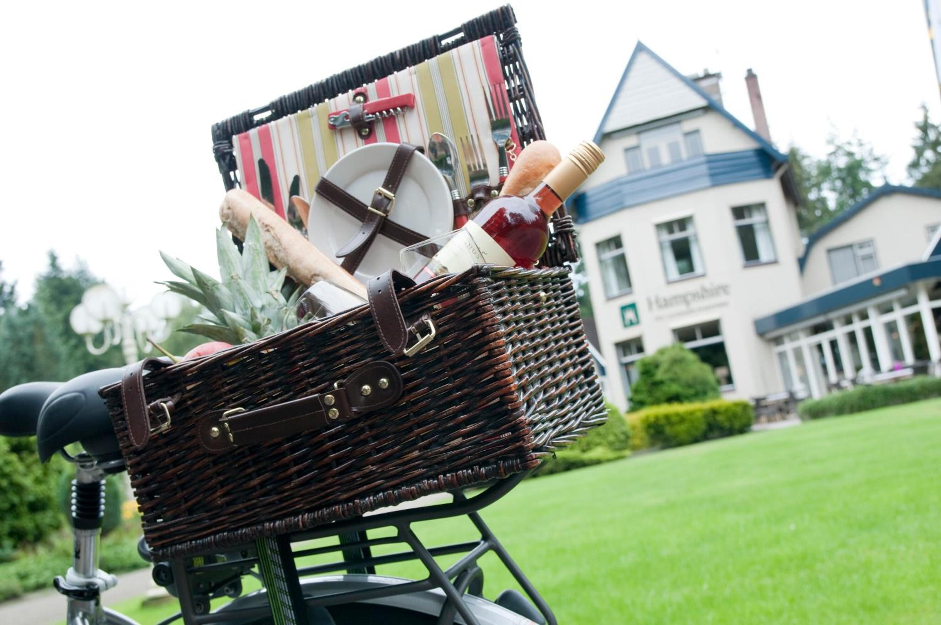 Un cesto di vimini su una bicicletta con una bottiglia, un piatto e un cavatappi. Sullo sfondo, un edificio con un cartello che recita 'Hampstead'.